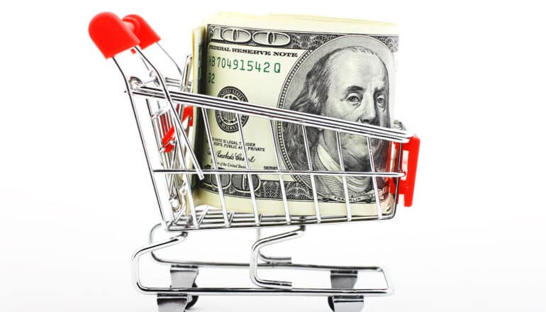 A miniature grocery cart with a hundred dollar bill in it against a white background.