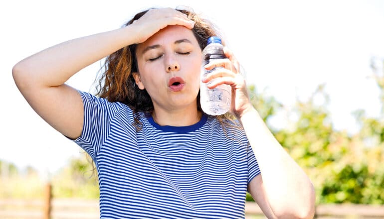 A young woman holds a water bottle to her face and a hand on her forehead while she sighs in hot weather.