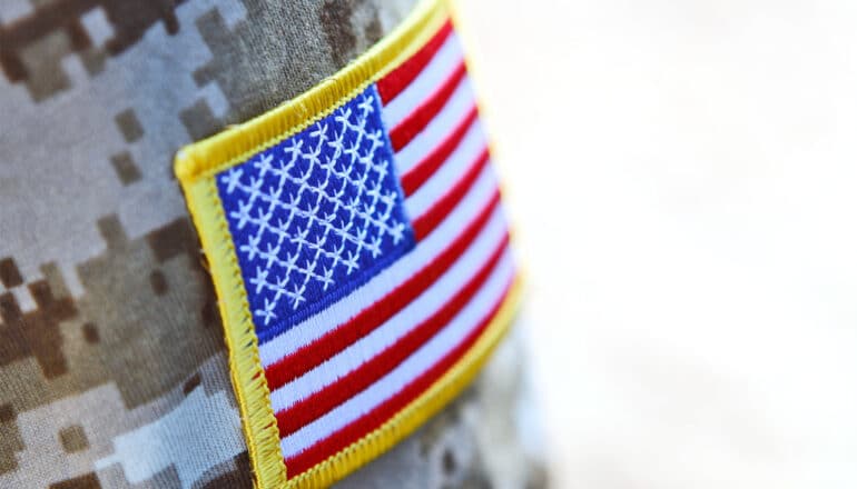 A soldier in camo uniform wears an American flag patch on their shoulder.