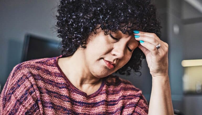 A woman puts her hand to her head and closes her eyes while seated in a living room.