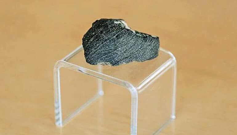 The meteorite, a small dark rock, sits on a small, clear plastic stand.