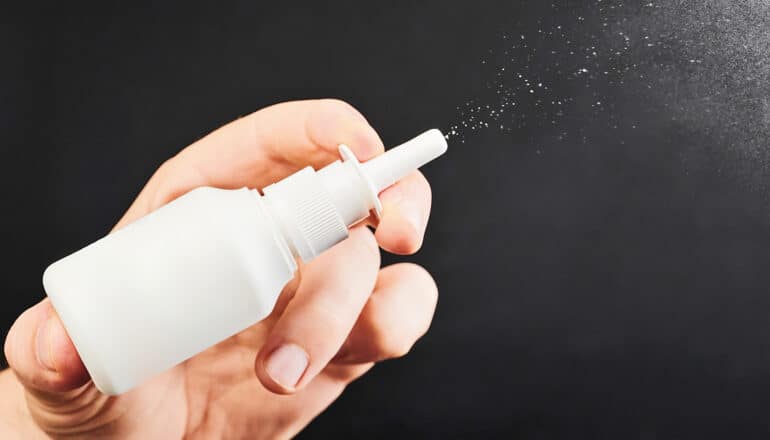 A person holds a white nasal spray bottle and sprays it against a dark background.