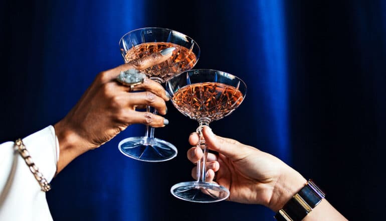 Two people toast with cocktail glasses filled with a red drink.