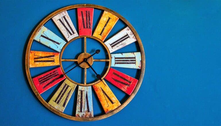 A colorful clock with Roman numerals hangs on a blue wall.