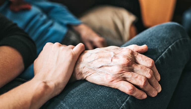 An older person rests her hand on a younger woman's leg while the younger woman holds her wrist.