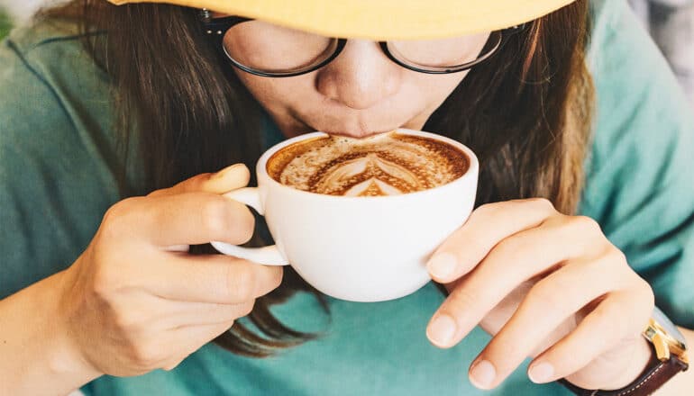 A young woman sips coffee from a white cup while leaning down.