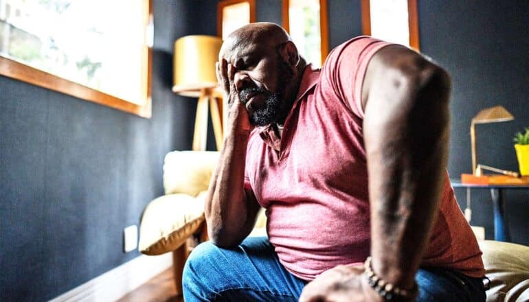 A man rubs his face with one hand while sitting on the edge of a bed looking stressed out.