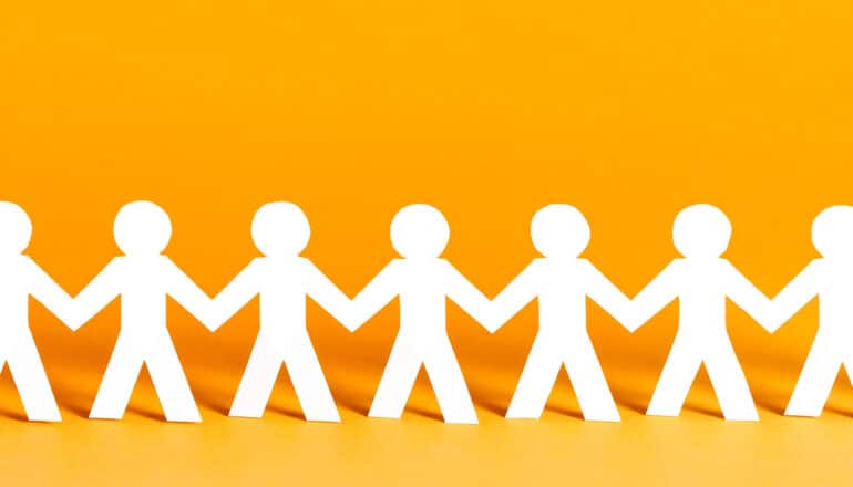 A chain of people cut out of white paper stands in front of a yellow background.