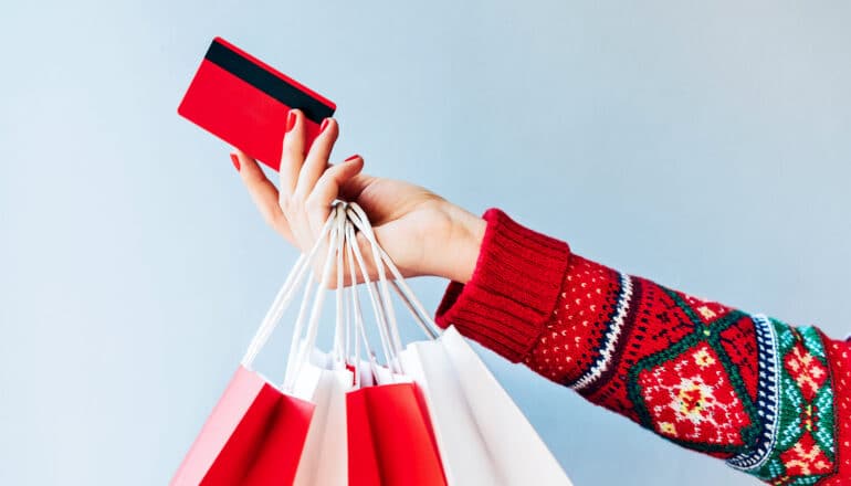 A woman wearing a holiday sweater holds out a credit card while shopping bags hang from her hand.