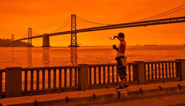 A person stands on the shore taking a picture of the Golden Gate Bridge while wildfire smoke turns the sky orange.