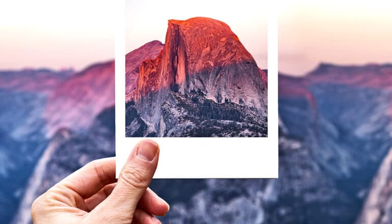 A person holds up a polaroid photograph showing a mountain in front of a wider mountain range.