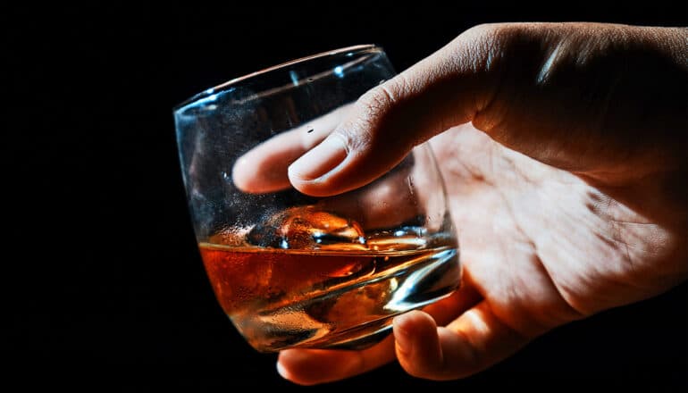 A person holds a glass of brown liquor against a dark background.