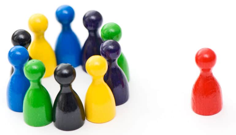 A group of colorful board game pieces stand in a circle while a single red game piece stands apart from the group.