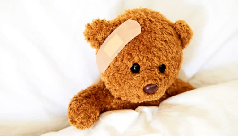 A teddy bear is tucked under a white blanket with a band-aid on its head.