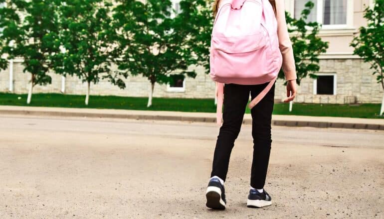 A young student walks to school on a tree-lined street while wearing a pink backpack.