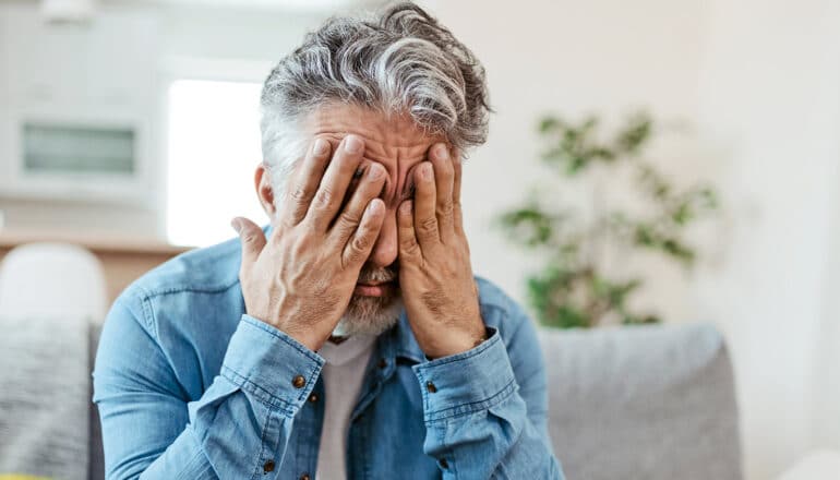 A man covers his face with both hands while sitting on a couch.