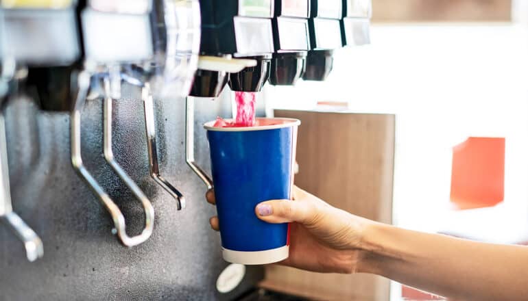 A person holds a cup under a soda fountain as it pours a pink drink.