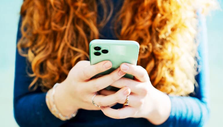 A redheaded woman holds her green cell phone in both hands.