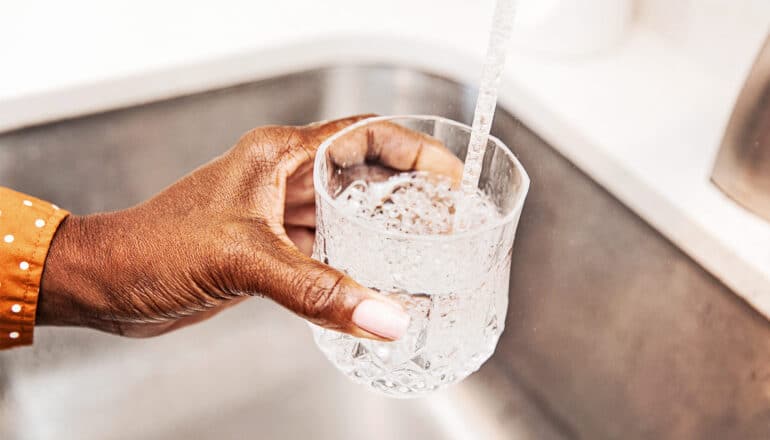 A person fills a glass with water from a kitchen sink tap.