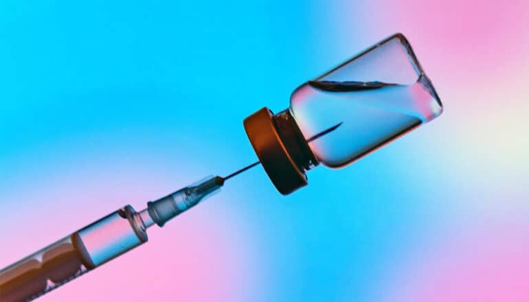 A syringe sticks into a vial filled with liquid while in front of a colorful blue, pink, and green background.