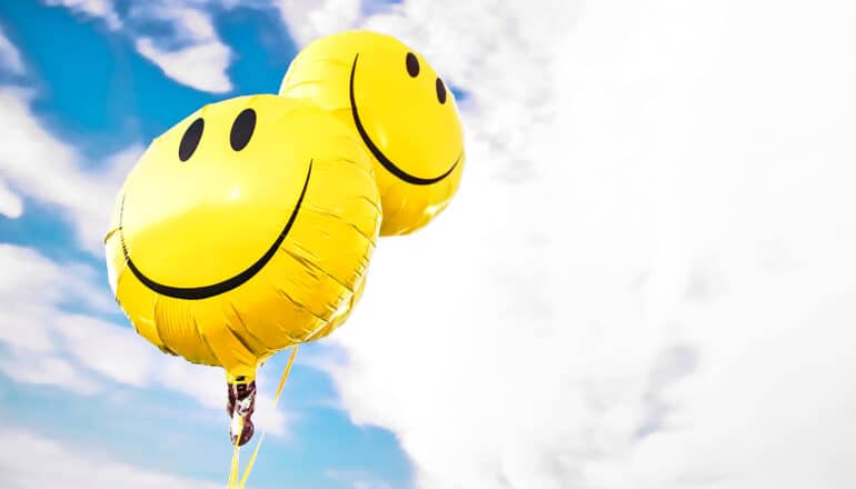 Two yellow balloons with smiley faces on them float against a blue sky with white clouds in it.