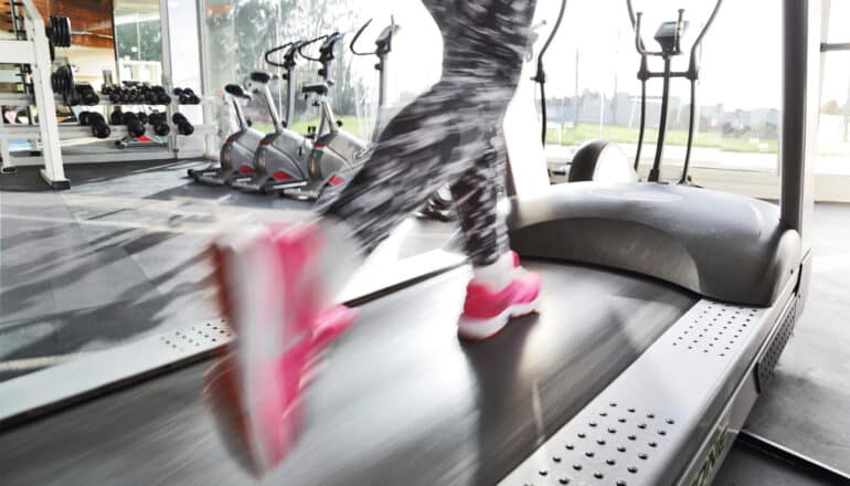 A person's legs blur as they're running on a treadmill in front of a gym window.