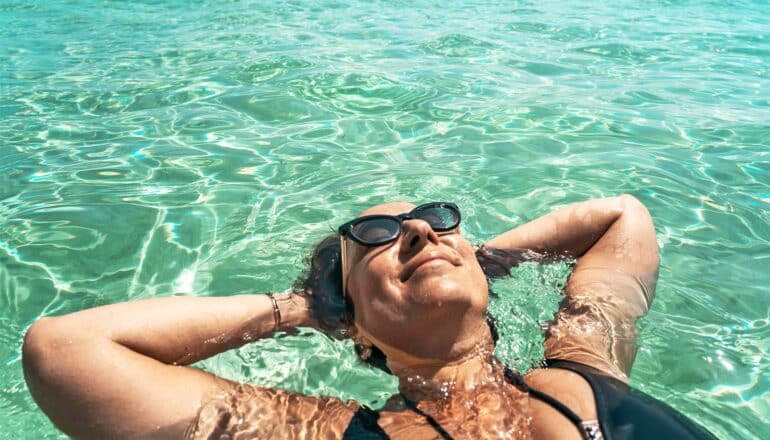 A woman floats on her back in a pool with her hands behind her head and a slight smile on her face.