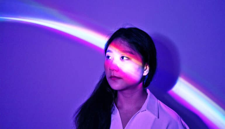 A young woman looks up to her right while a colorful beam of light shines across her face.