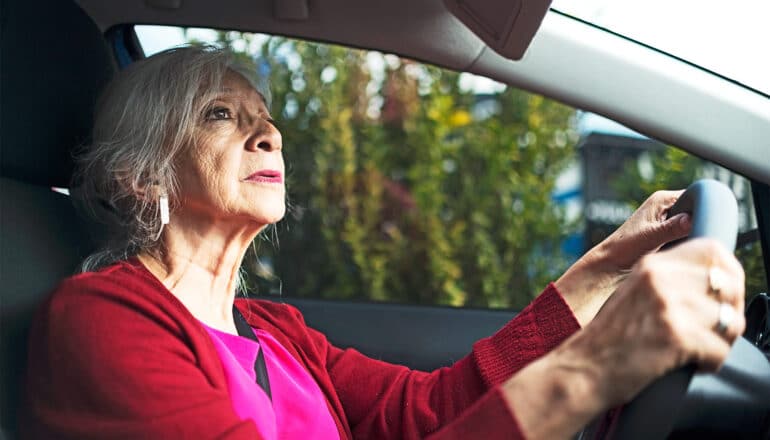 An older woman wearing red looks in the rear view mirror while driving.