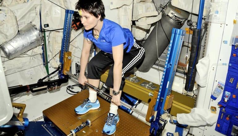 An astronaut in workout gear lifts a weighted bar while on the ISS.