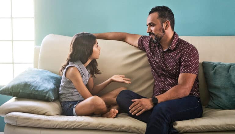 A father and daughter sit and talk on a couch in front of a blue wall.