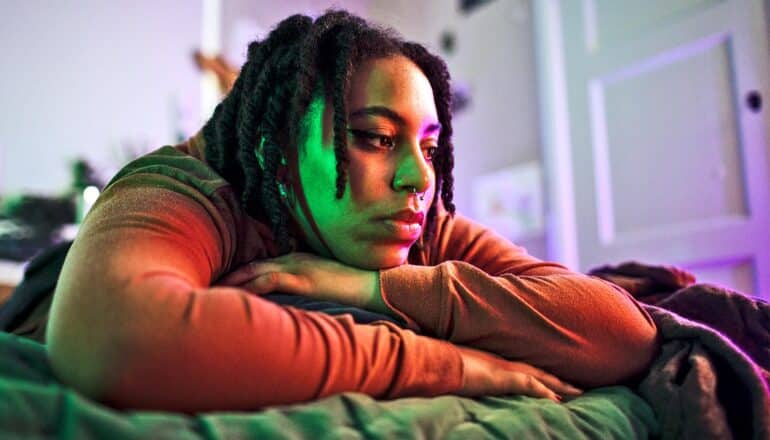 A young woman lays on her stomach on a bed with a blank expression on her face.