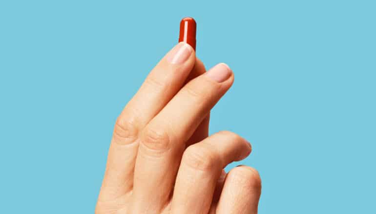 A person holds a single red pill in their fingers against a blue background.