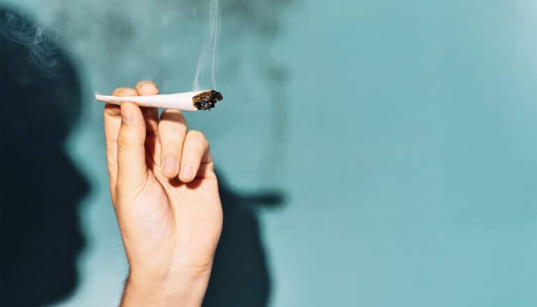 A person holds a smoking joint in one hand in front of a blue background.