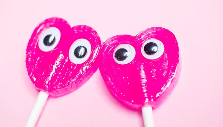 Two pink heart-shaped lollipops with googly eyes attached to them.