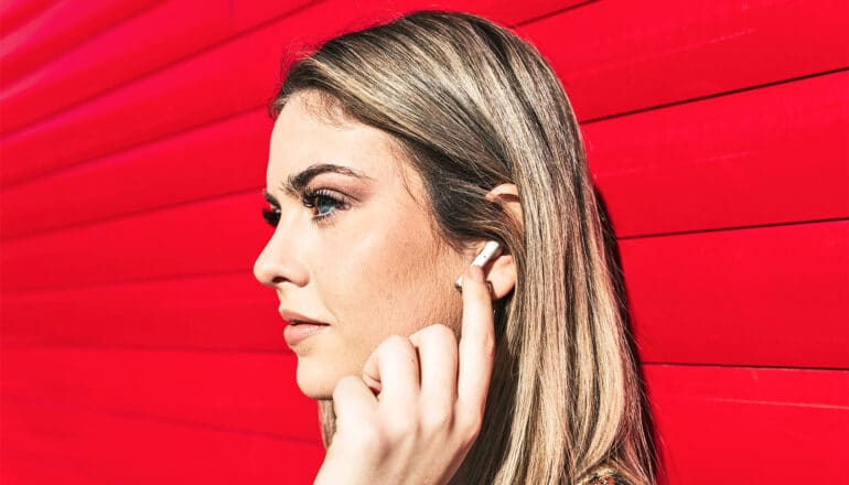 A young woman taps the white wireless headphone in her ear while leaning against a red wall.