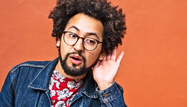 A man wearing a jean jacket and glasses leans to one side and holds his hand to his ear to hear.