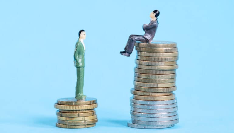 One small figurine of a person in a suit stands on a short stack of coins while another sits on a taller stack of coins.