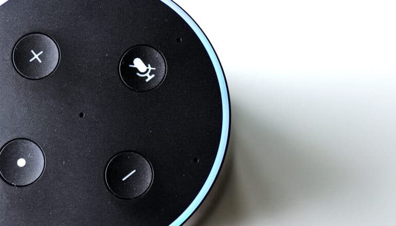 A close-up shot of the top of a smart speaker shows several buttons, including volume up and down and a voice command button.