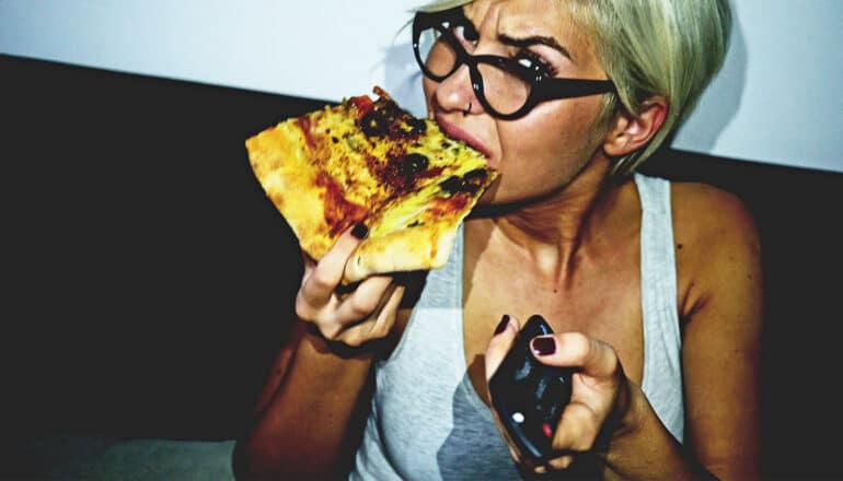 A woman wearing glasses eats a piece of pizza while holding a remote and making an annoyed face.