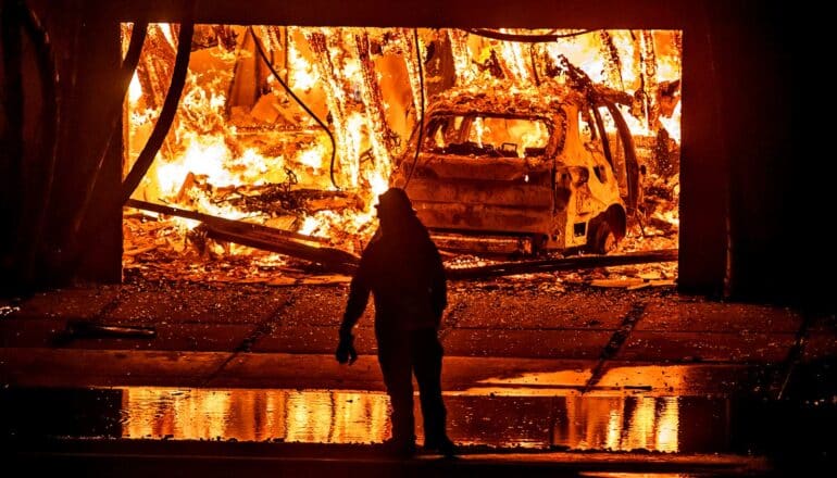 A firefighter looks into a home's garage as fire engulfs a car and the entire home's interior.