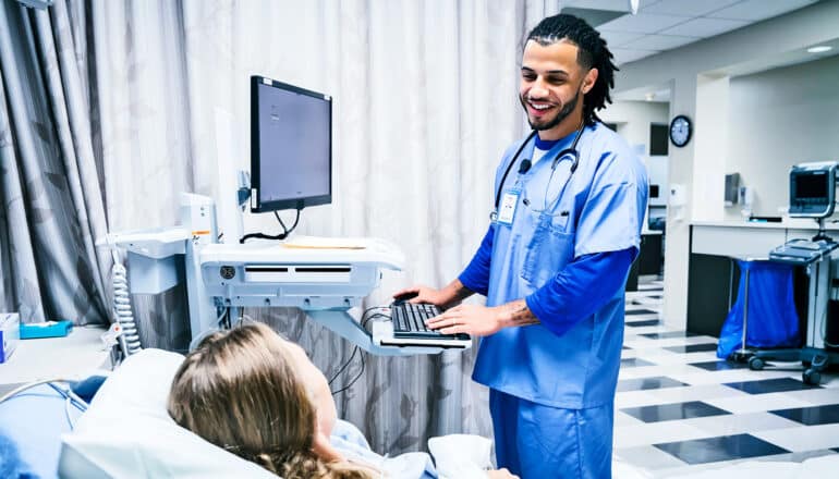 A nurse stands at a computer station at a patient bedside and smiles while he talks to the patient.