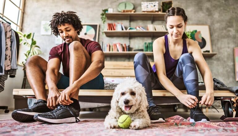 A man and woman lace up running shoes while their dog sits between them.