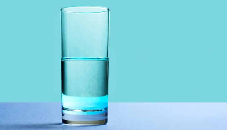 A glass of water that's half full or half empty sitting on a blue surface in front of a blue background.