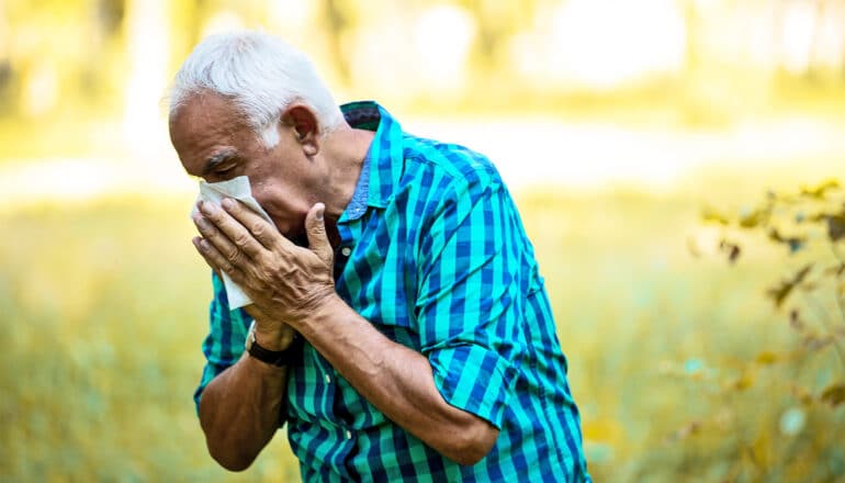 An older man sneezes into a tissue while walking outside on a sunny day.