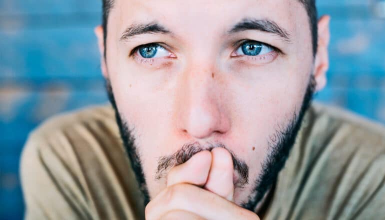 A young man with blue eyes puts his hands to his lips and looks stressed or lost in thought.