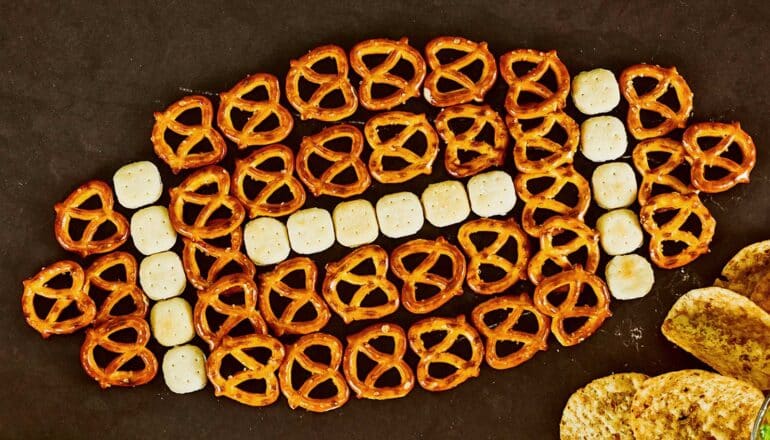 Pretzels and oyster crackers arranged in the shape of a football on a dark surface.