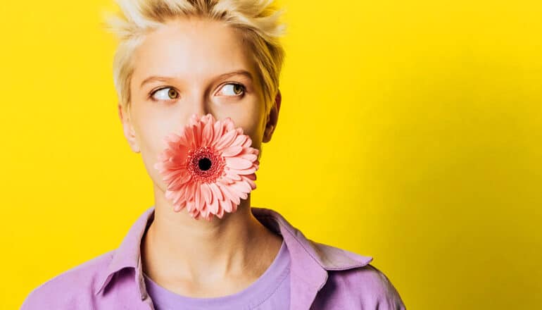 A woman holds a pink flower in her mouth as she stands in front of a yellow background.