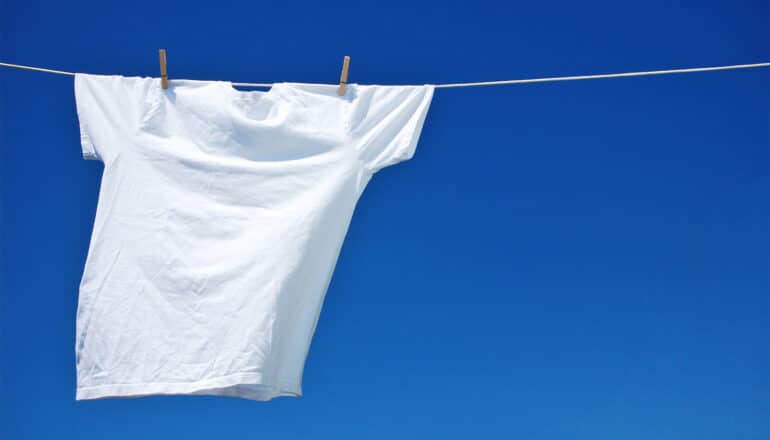 A white t-shirt air dries while hanging on a clothing line in front of a blue sky.