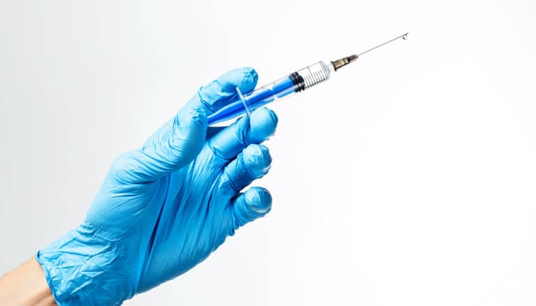 A person wearing a blue glove holds a syringe against a white background.